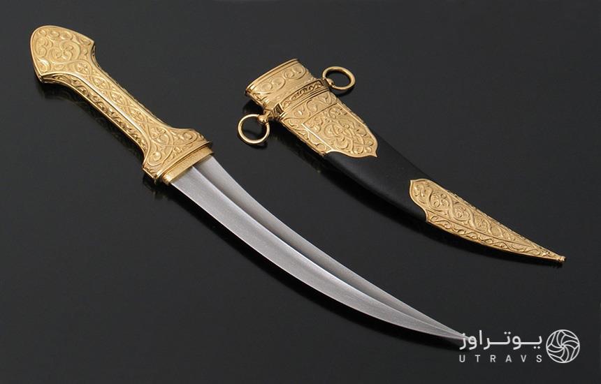 خنجری با دسته‌ی طلایی روی پس‌زمینه‌ای سیاه‌رنگ و غلاف طلایی‌-مشکی خنجر در کنارش